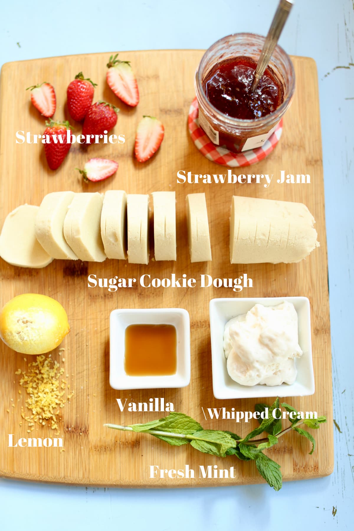 a wooden cutting board with recipe ingredients on top, captioned...cookie dough, strawberries, whipped cream, lemon and strawberry jam.