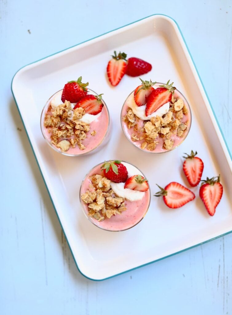 three strawberry shortcake smoothies on a square white platter with fresh srwaberry garnish on a blue table.