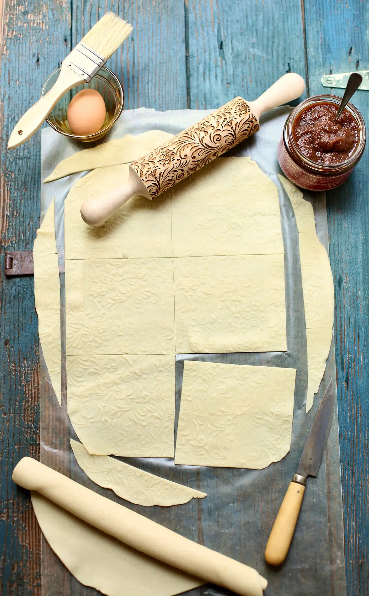 a sheet of pastry dough on a blue wooden board with a rolling pin, an egg, knife and jar of apple butter.