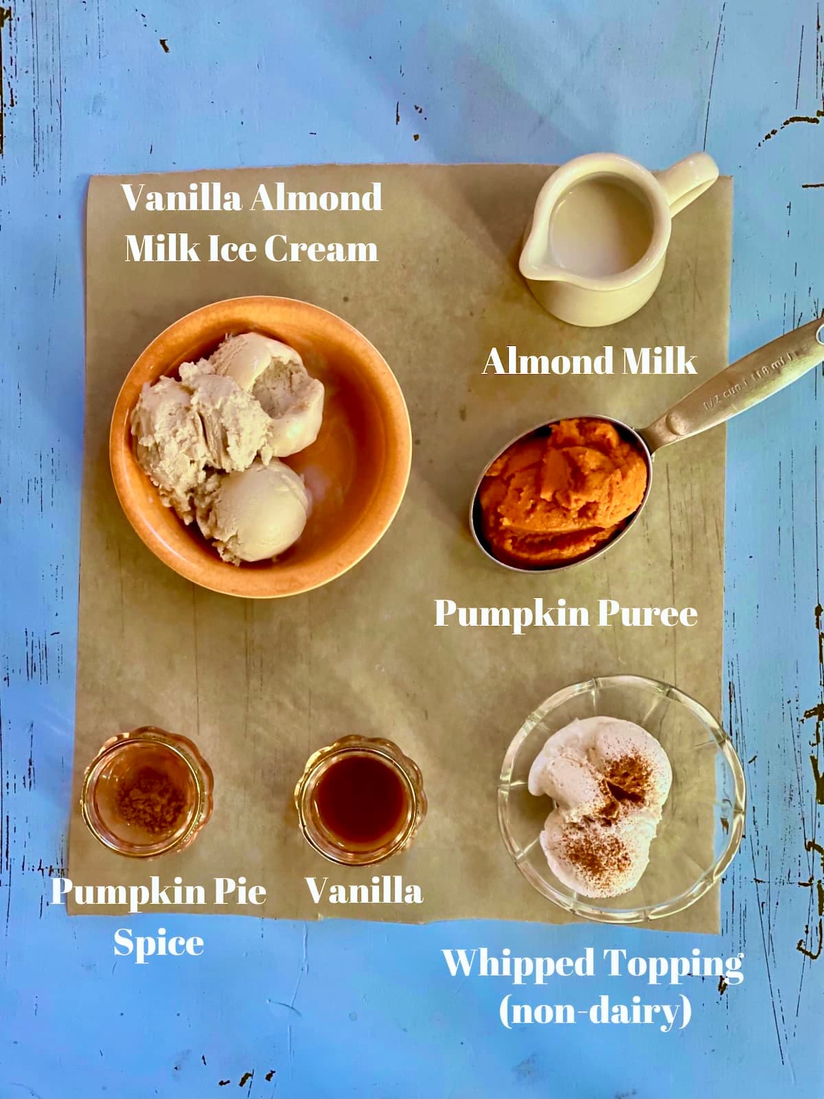 a blue table with ingredients, vanilla ice cream, pumpkin puree, almond milk, pumpkin pie spice, vanilla, with a text overlay saying those ingredient names.
