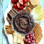 a dessert tray with chocolate dip and cookies and fruit on a white platter with a light blue background.