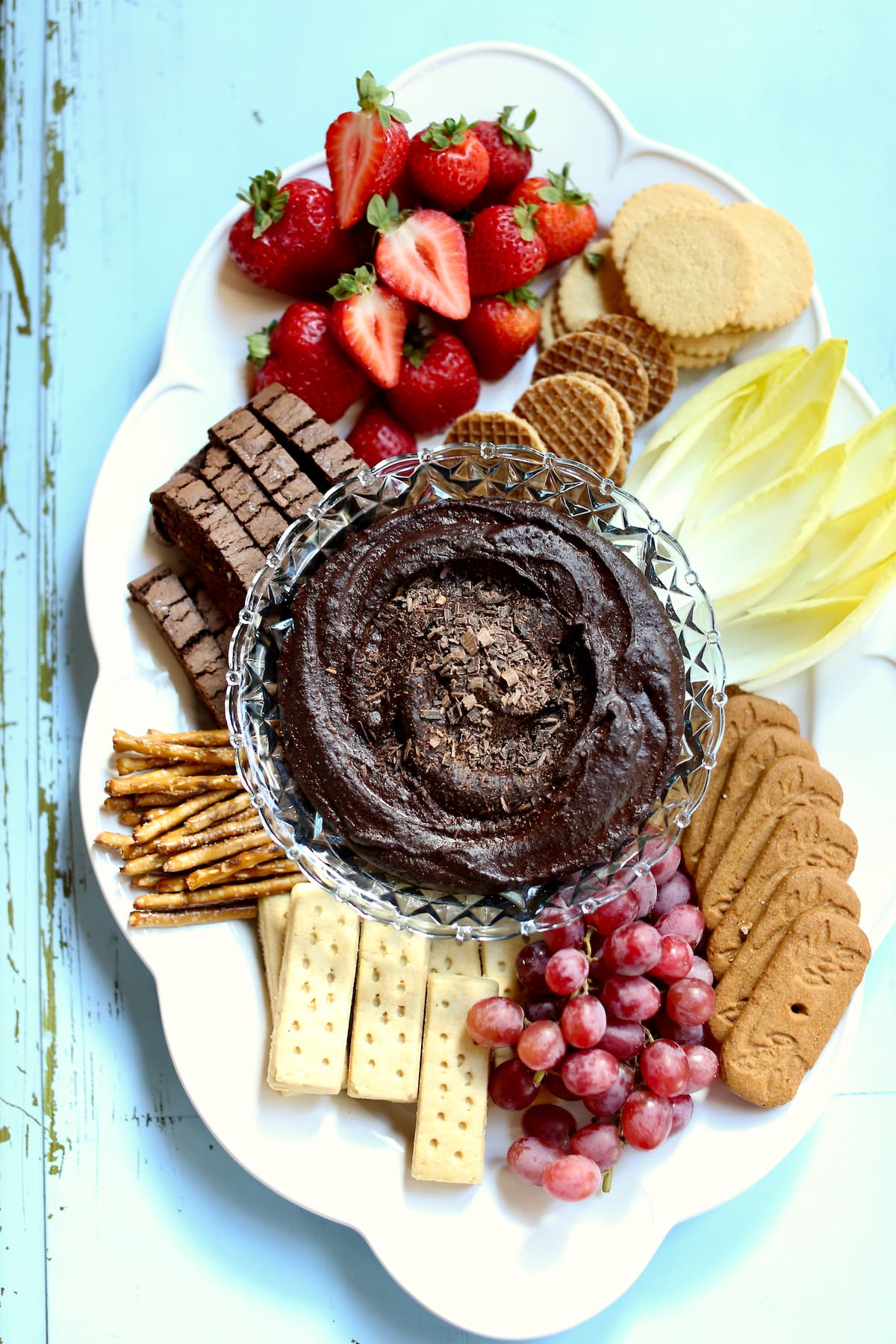 a dessert tray with chocolate dip and cookies and fruit on a white platter with a light blue background.
