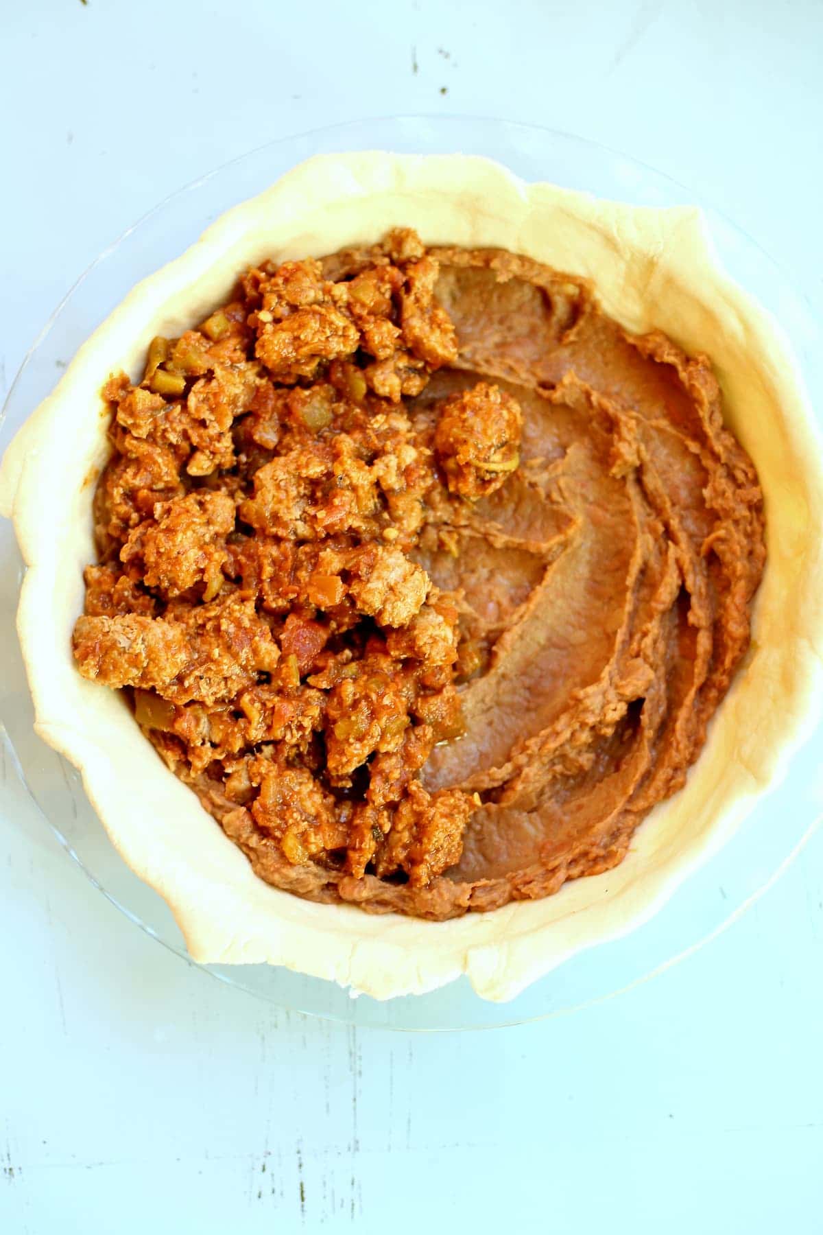 an uncooked meat pie with beans on one side and taclo meat on the other, sitting on a blue table.