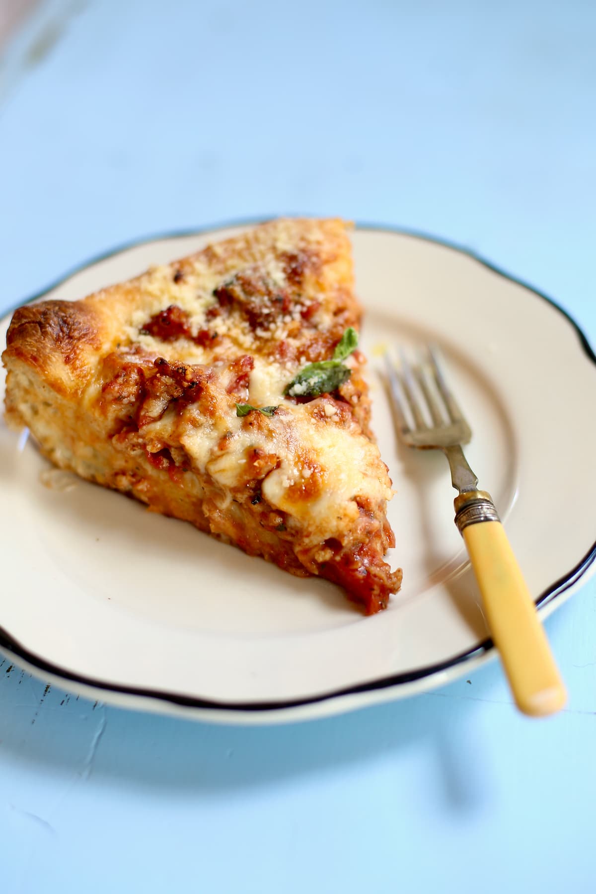 a piece of deep dish pizza on a white plate with a fork on a blue table.