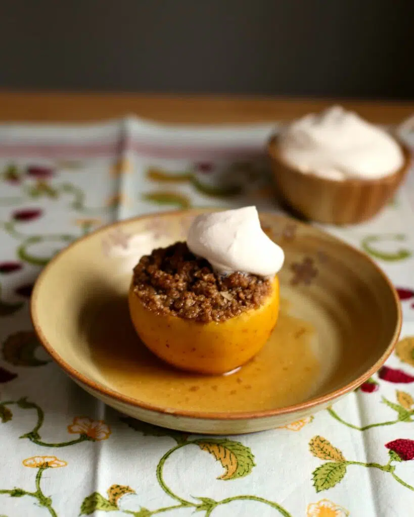 one baked apple with whipped cream on top, and whipped cream in the background, on a table that has a tablecloth on it.