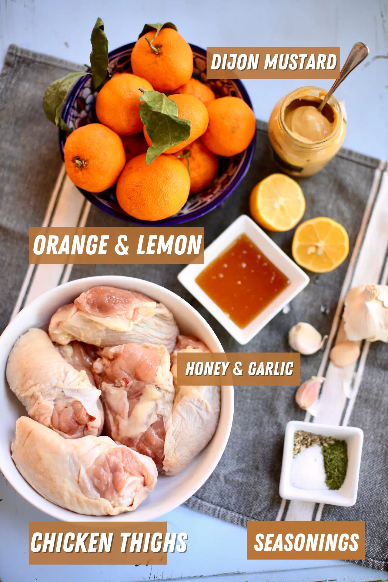 a gray tablecloth is behind several ingredients: oranges and lemons, chicken thighs, honey, garlic and seasonings.