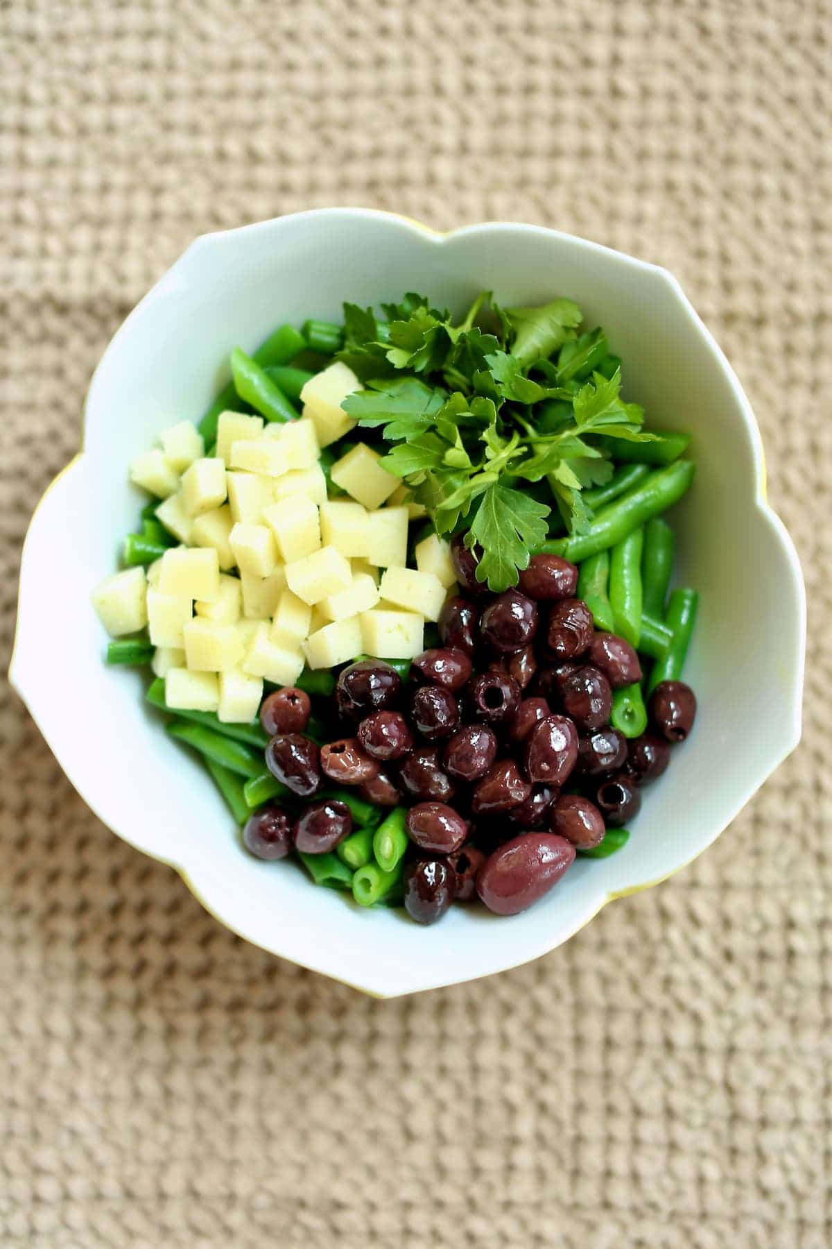 a white bowl with cubed cheese, kalamata olives, green beans and more on a table.