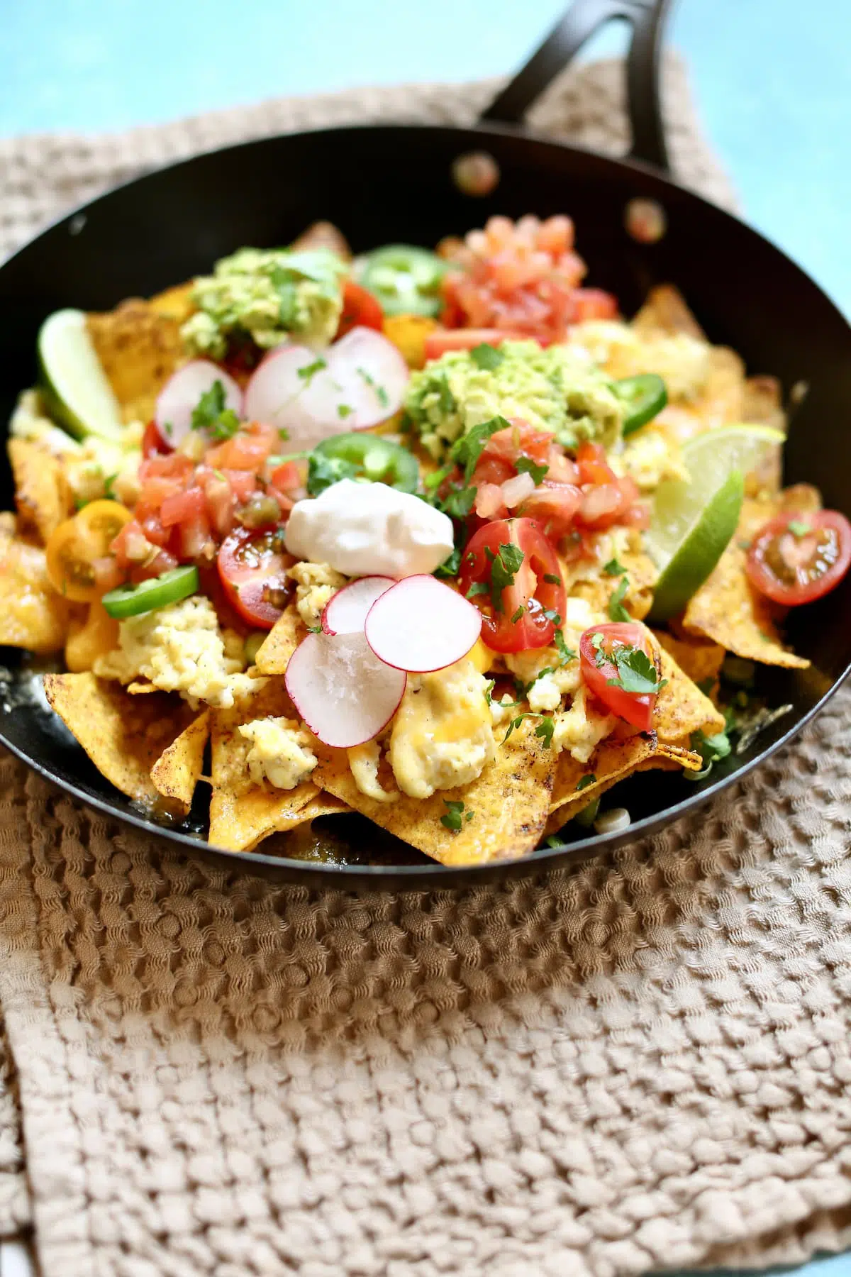 a skillet of nachos with sour cream, green onions, cheddar cheese and eggs on a table.