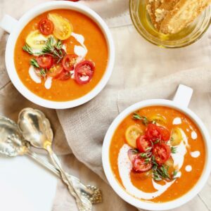 two bowls of tomato soup and spoons on a napkin with crackers.