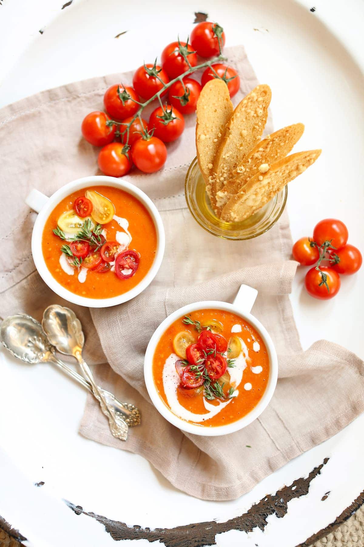 a white platter with two white bowls of tomato soup and some crackers on them, along with some fresh tomatoes and spoons.