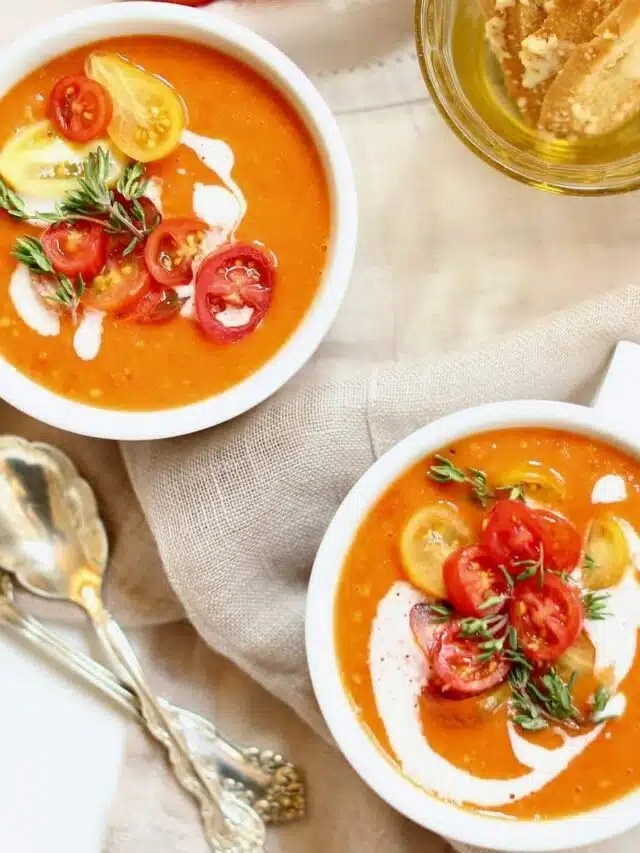 two bowls of tomato soup and spoons on a napkin with crackers.