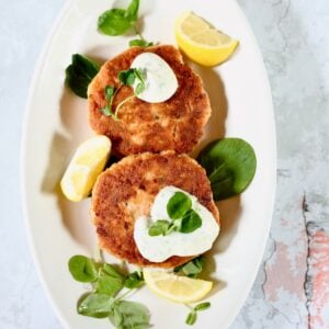 a square photo of two salmon patties on a platter with fresh dill sauce.