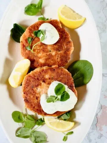 a square photo of two salmon patties on a platter with fresh dill sauce.