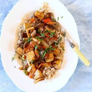 a square photo of eggplant tofu on a white plate and blue table.