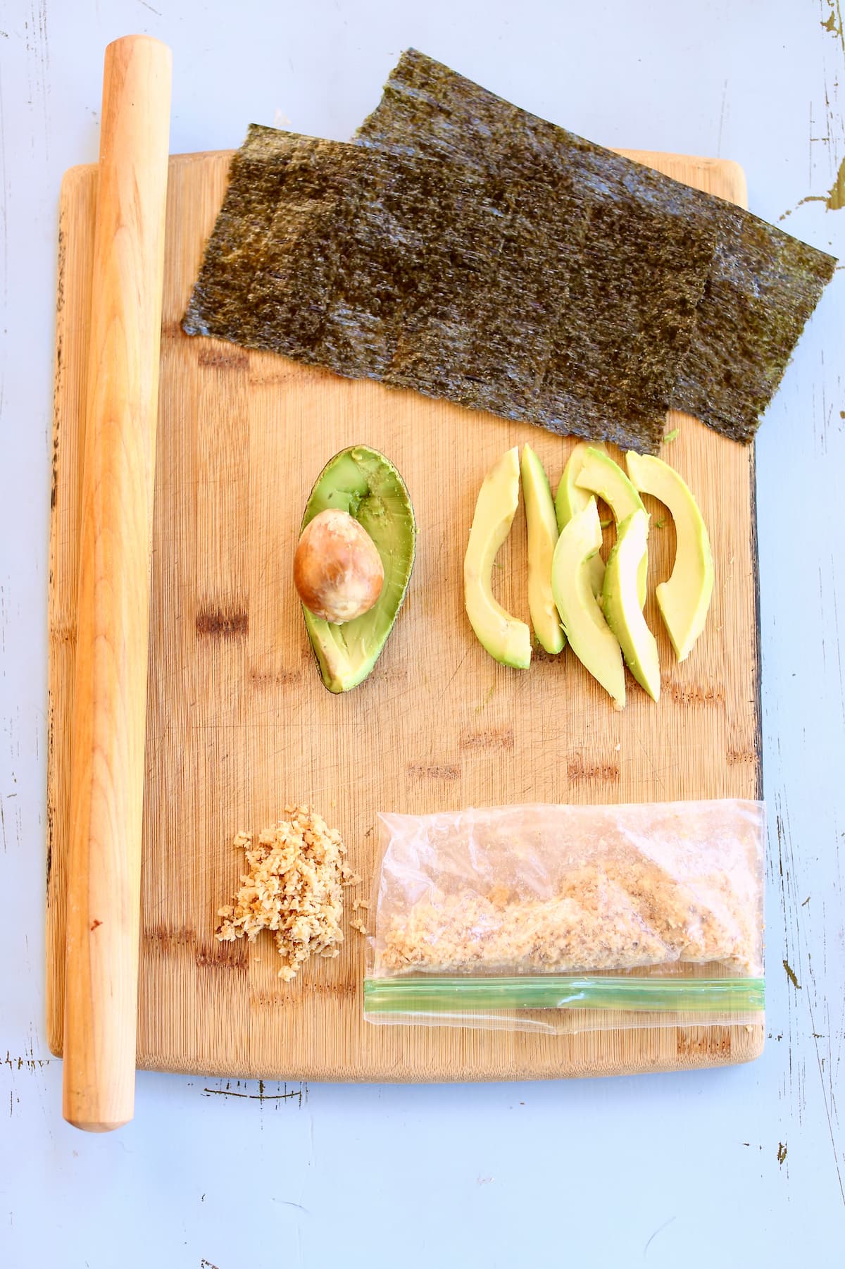 Nori sheets, avocado, and other ingredients for a sushi roll on a cutting board.