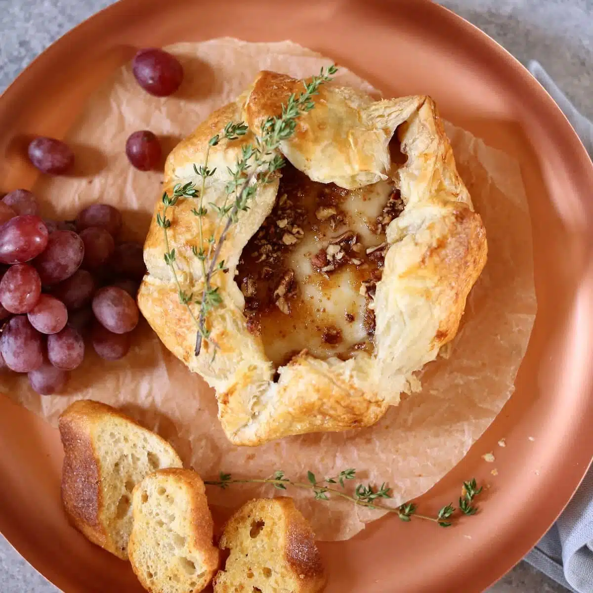 a square photo of a brie in puff pastry o a copper tray with grapes.