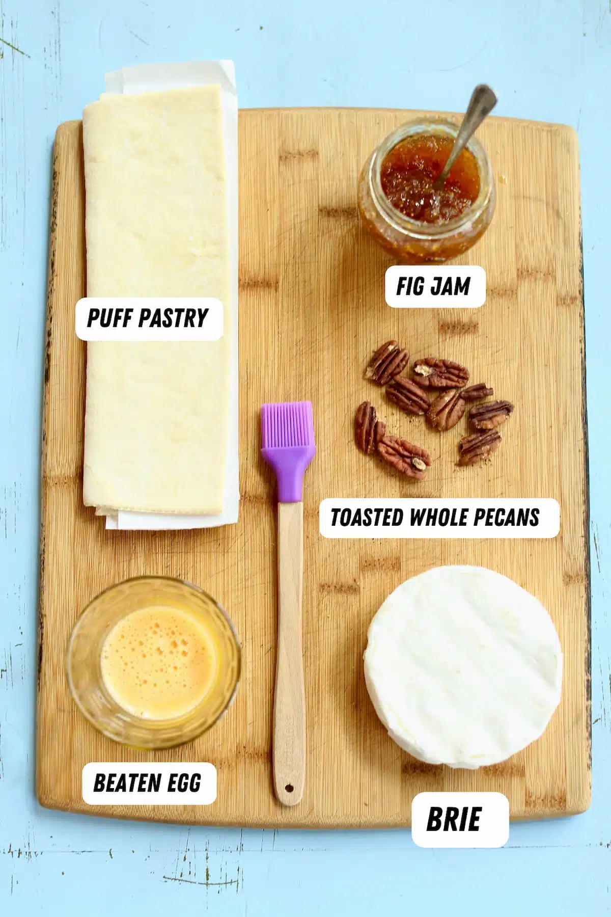 a cutting board of a wheel of brie cheese, egg wash, pecans, fig jam and puff pastry.