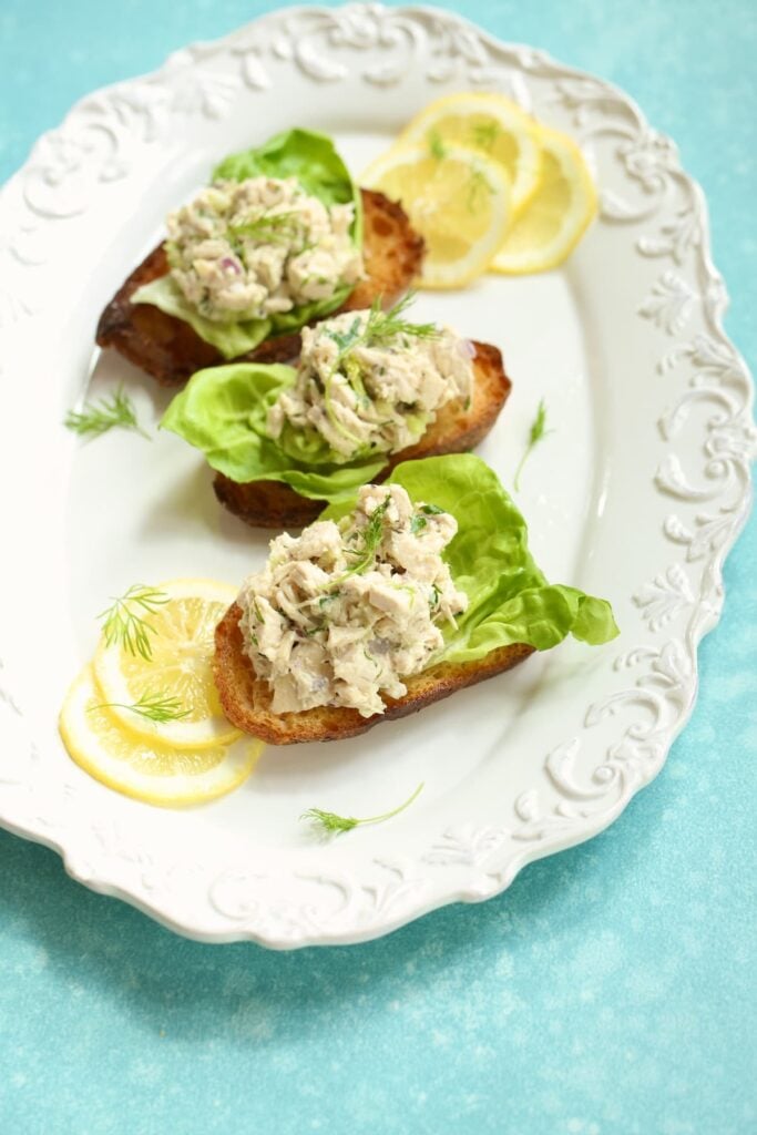 A white platter with three classic savory chicken salad sandwiches, with fresh lemon garnish.