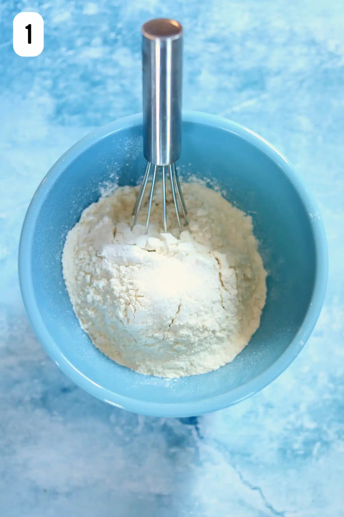 A blue bowl of dry ingredients for cupcakes with a whisk coming out of the side.