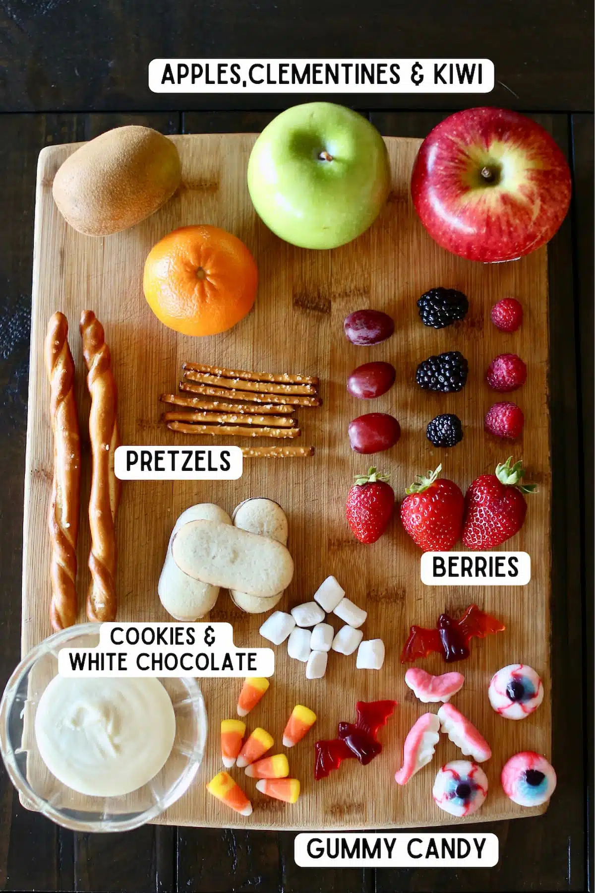 a cutting board of ingredients, candy and fruit, to make fruit halloween snacks.